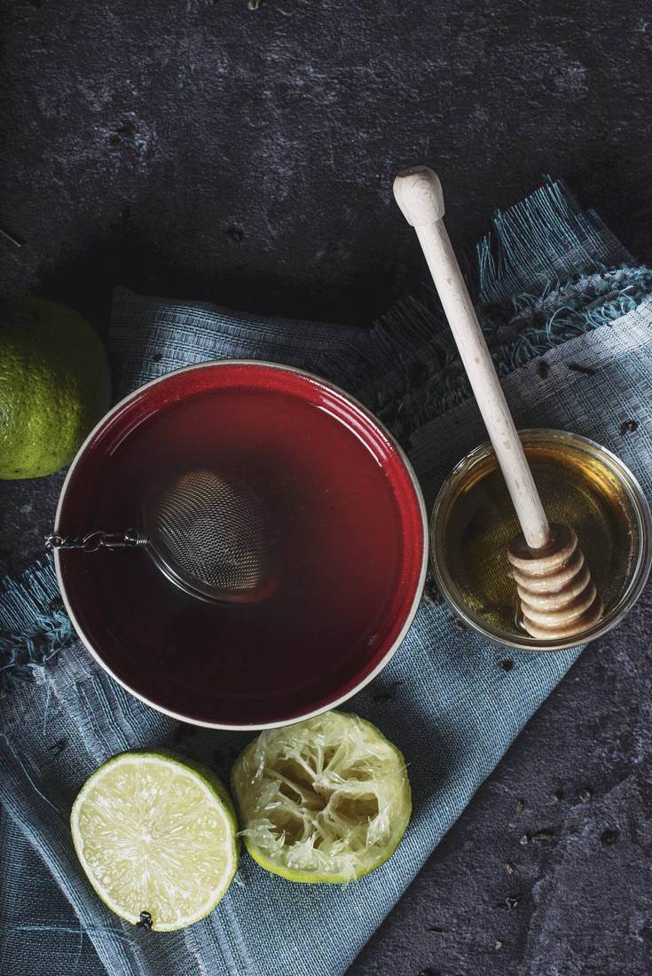 A cup of ruby-red rose hibiscus tea on dark slate, with honey and lime